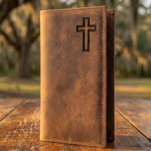 May include: A brown leather wallet with a cross design. The wallet is made of a distressed leather material and has a vertical orientation. The cross is etched into the front of the wallet. The wallet is open, revealing the interior.
