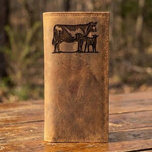 May include: A brown leather wallet with a stitched border. The front of the wallet features a detailed engraving of a cow and two calves. The wallet is rectangular and appears to be made of distressed leather.