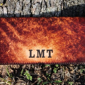 May include: A brown leather wallet with the initials "LMT" embossed on the front. The wallet has a textured surface and a stitched border. The background is a tree trunk and some grass.