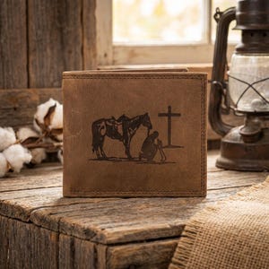 May include: Brown leather wallet with a detailed image of a horse, a person kneeling, and a cross. The wallet is rectangular and appears to be bi-fold. The image is etched into the leather. The wallet is sitting on a wooden surface.