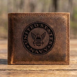 May include: Brown leather wallet with the United States Navy emblem embossed on the front. The emblem features an eagle, anchor, and text that reads "United States Navy" around the border. The wallet is rectangular and appears to be new.