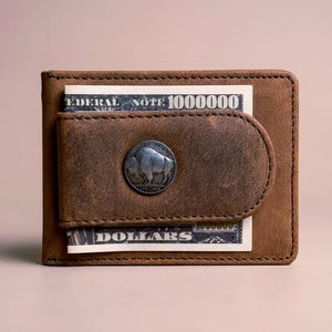 May include: A brown leather wallet with a metal money clip. The clip features a buffalo design. The wallet holds a Federal Note and a dollar bill.
