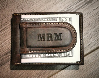 May include: A brown leather wallet with a money clip and the initials "MRM" embossed on the front. The wallet is closed and has a bill sticking out of the top.