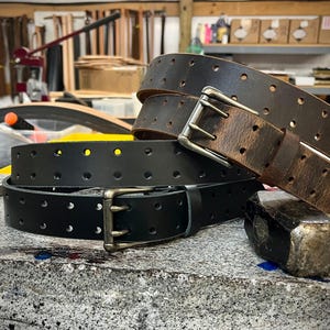 May include: Three leather belts in varying shades of brown and black, each with a metal buckle. The belts are perforated with evenly spaced holes. A hammer and other tools are visible in the background, suggesting a workshop setting.