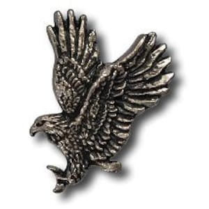 May include: A detailed, silver-colored metal pin in the shape of an eagle in flight. The pin features intricate feather detailing and a sharp beak, with outstretched wings. The eagle is depicted mid-flight, with its talons extended.