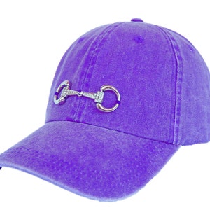 May include: A purple baseball cap with a silver metal equestrian bit detail. The bit has a rhinestone design.