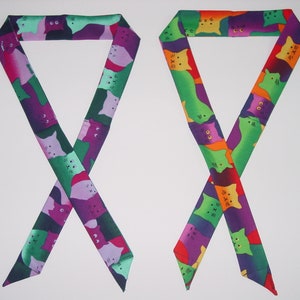 Op de afbeelding: Twee kleurrijke stoffen haarbandjes met een kattenprint. De bandjes zijn groen, paars, oranje en roze met een witte achtergrond. De katten staan in verschillende poses en hebben verschillende gezichtsuitdrukkingen.