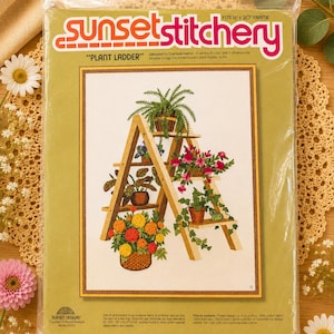 Könnte beinhalten: Ein Sunset Stitchery Stickset mit dem Titel Plant Ladder in einer Kunststoffverpackung. Das Cover zeigt ein gedrucktes Motiv eines hölzernen A-förmigen Pflanzenständers mit verschiedenen Topfpflanzen und Blumen. Passend für einen 40,6 cm x 50,8 cm Rahmen.