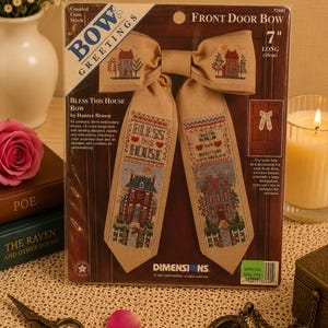 May include: A Dimensions counted cross stitch kit for a 7 inch long Front Door Bow by Hunter Brown. The beige fabric features embroidered houses, floral motifs, and the text Bless This House. The packaging includes instructions and a chart.