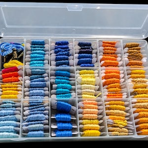 May include: A clear plastic storage box containing multiple compartments filled with embroidery floss wound onto white cardboard bobbins. The collection features a variety of colors including shades of blue, yellow, orange, and gold.