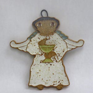 May include: A ceramic angel ornament with a white robe, holding a green chalice. The angel has a brown face, blue accents, and a black loop for hanging. The ornament has a speckled texture.