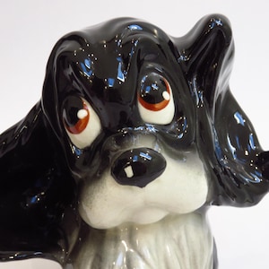 May include: A ceramic dog figurine with glossy black fur and large, expressive eyes. The dog has a white muzzle and chest, with a black nose and floppy ears. The figurine is a decorative item, likely for display.