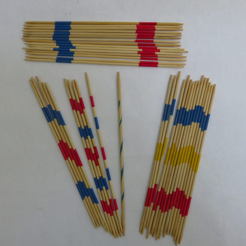 May include: A collection of wooden Mikado game sticks. The sticks are natural wood color with painted bands of red, blue, and yellow. Some sticks are bundled, others are fanned out, and one is isolated.