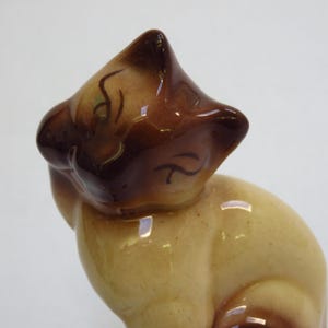 May include: A ceramic figurine of a Siamese cat, featuring a glossy brown head and ears, with a cream-colored body. The cat is in a seated position, with its head tilted slightly to the side. The figurine has a vintage aesthetic.
