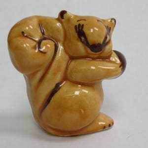 May include: A ceramic squirrel figurine in a curled pose, holding a nut. The squirrel is a light brown color with darker brown accents and facial features. The figurine has a glossy finish and is set against a plain background.