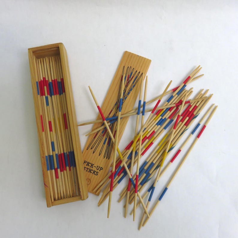 May include: A wooden pick-up sticks game set. The set includes a rectangular wooden box, a wooden board with black lines, and numerous wooden sticks with red, blue, and yellow painted tips. The words "PICK-UP STICKS" are printed on the board.