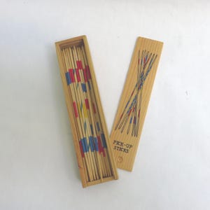 May include: Wooden pick-up sticks game with a rectangular box and lid. The box contains numerous wooden sticks with painted red, blue, and yellow sections. The lid is decorated with a design of crossed sticks and the words "PICK-UP STICKS".