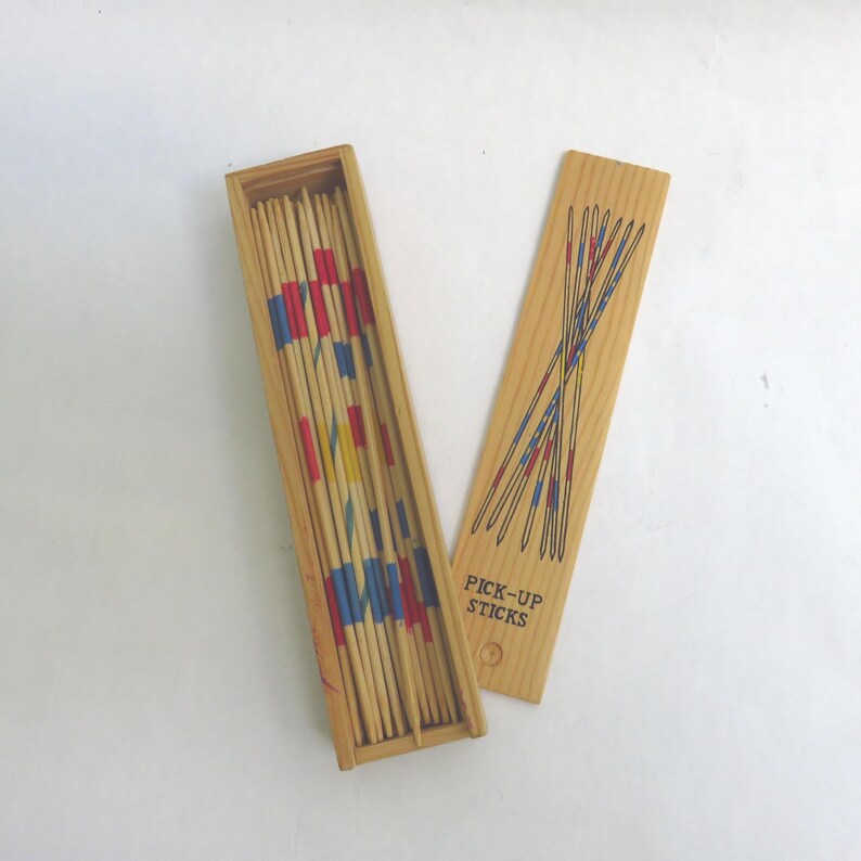 May include: Wooden pick-up sticks game with a rectangular box and lid. The box contains numerous wooden sticks with painted red, blue, and yellow sections. The lid is decorated with a design of crossed sticks and the words "PICK-UP STICKS".