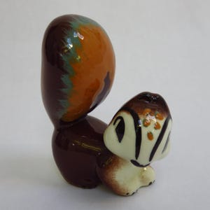 May include: A ceramic squirrel figurine with a glossy finish. The squirrel has a large, rounded tail in shades of brown, orange, and teal. The head and body are a mix of brown, cream, and black. The figurine is a decorative item.
