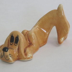 May include: A ceramic figurine of a cartoon dog in a lying down position. The dog is light brown with black eyes, a black nose, and a smiling mouth. The tail is curved upwards. The figurine is on a white surface.