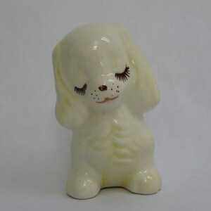 May include: A cream-colored ceramic puppy figurine with closed eyes and long eyelashes. The puppy has a small brown nose and mouth, and its ears are drooping. The figurine has a glossy finish and is standing on all fours.