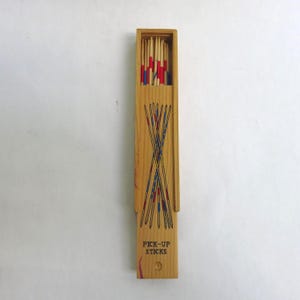 May include: Wooden Pick-Up Sticks game set. The rectangular wooden box is open, revealing a collection of colorful sticks. The box is light brown with a design of crossed sticks and the words "PICK-UP STICKS" printed on the front.
