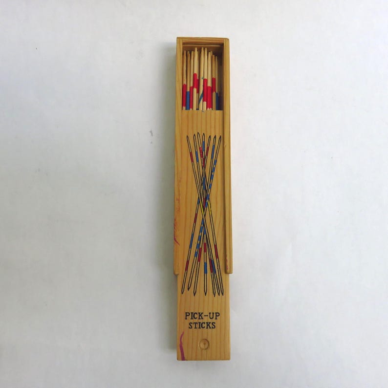 May include: Wooden Pick-Up Sticks game set. The rectangular wooden box is open, revealing a collection of colorful sticks. The box is light brown with a design of crossed sticks and the words "PICK-UP STICKS" printed on the front.