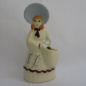 May include: A ceramic figurine of a person wearing a wide-brimmed light blue hat, a cream-colored dress with light blue polka dots, and a dark brown bow at the neck. The figure holds a hollow, cream-colored basket-shaped container on the right side.