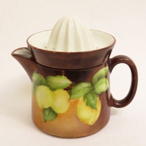 May include: A ceramic citrus juicer with a brown base and a white reamer. The juicer features a spout and a handle. The base is decorated with a painted design of lemons and green leaves.