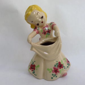May include: A vintage ceramic figurine of a girl in a dress, with yellow hair and closed eyes. The dress is beige with red and green floral accents. The figurine is holding up the bottom of her dress, revealing a vase opening.