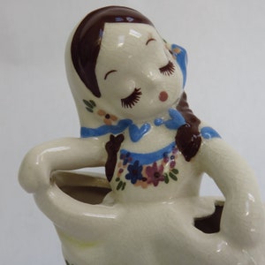 May include: A vintage ceramic figurine of a woman with closed eyes, brown hair, and a cream-colored dress. The dress is adorned with blue and purple floral patterns and a blue ribbon. The figurine has a glossy finish.