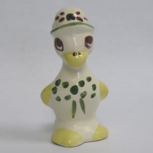 May include: A ceramic duck figurine with a cream-colored body, yellow feet and wings, and a yellow beak. The duck wears a hat with brown spots and green trim. Green and brown floral accents decorate the front.