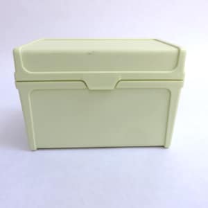 May include: A rectangular, pale yellow plastic storage box with a hinged lid. The container features a smooth, solid-colored surface and a simple latch mechanism centered on the front panel, designed for holding small items securely inside.