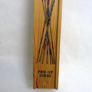 May include: Wooden box of Pick-Up Sticks game. The sticks are black with red, blue, and yellow accents. The words "PICK-UP STICKS" are printed in black on the box. The box is made of light-colored wood.