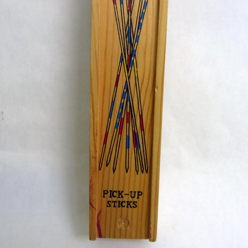 May include: Wooden box of Pick-Up Sticks game. The sticks are black with red, blue, and yellow accents. The words "PICK-UP STICKS" are printed in black on the box. The box is made of light-colored wood.