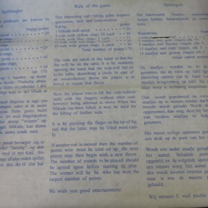 May include: A page from a game rulebook, featuring text in multiple languages. The text describes the rules of a rod-lifting game, including scoring and gameplay instructions. The paper has a slightly aged appearance.