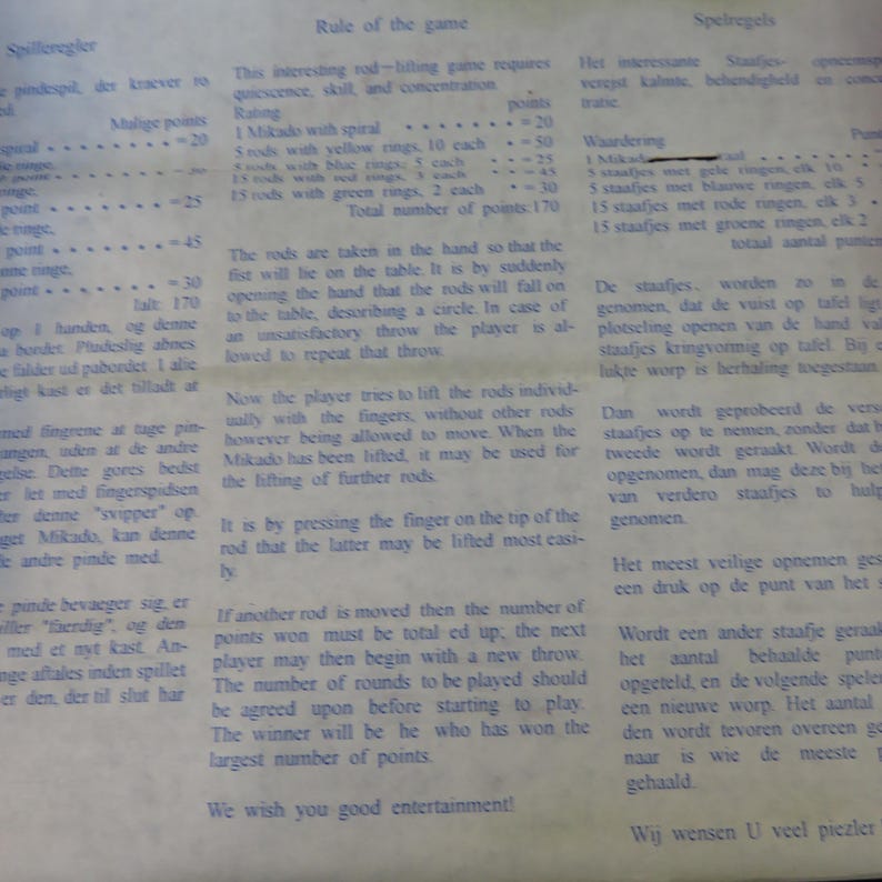 May include: A page from a game rulebook, featuring text in multiple languages. The text describes the rules of a rod-lifting game, including scoring and gameplay instructions. The paper has a slightly aged appearance.