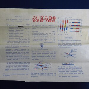 May include: A vintage Mikado Pick-Up Sticks game instruction sheet. The paper is aged, with text in multiple languages and illustrations of the game. The game includes sticks with colored stripes and point values.