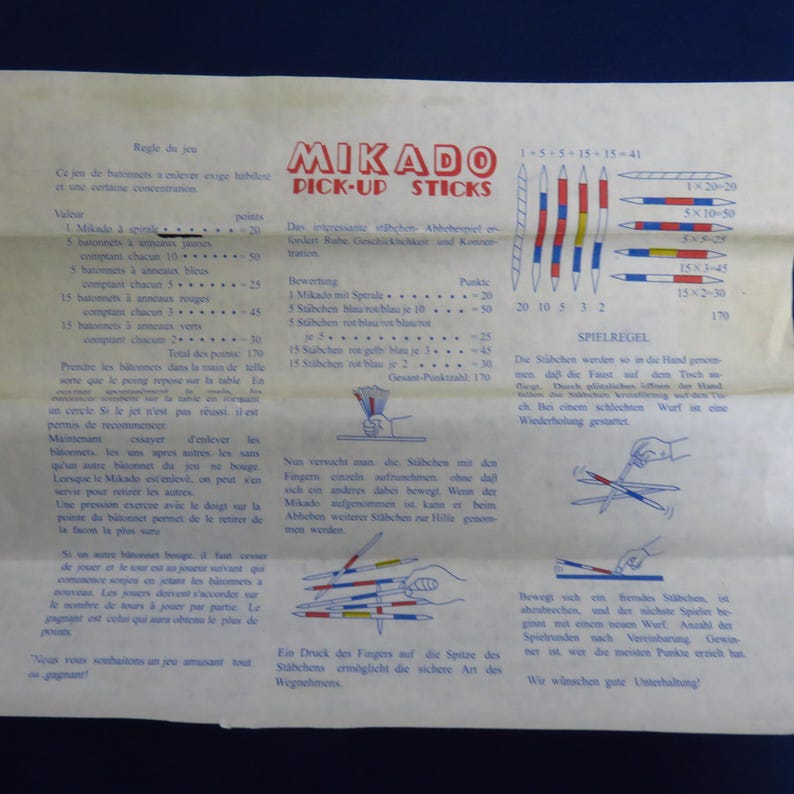 May include: A vintage Mikado Pick-Up Sticks game instruction sheet. The paper is aged, with text in multiple languages and illustrations of the game. The game includes sticks with colored stripes and point values.