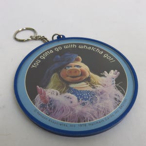 May include: A blue and white Miss Piggy keychain. The circular charm features Miss Piggy in a blue dress and feather boa, with the text "You gotta go with whatcha got!". The keychain is from 1978.