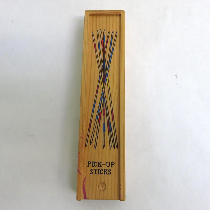 May include: Wooden pick-up sticks game in a rectangular box. The box is light brown with the words "PICK-UP STICKS" printed in black. The sticks are black with red and blue accents.