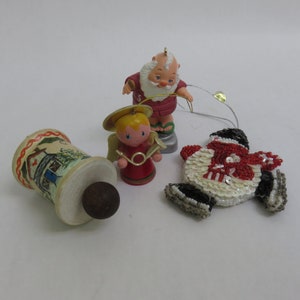 May include: A collection of four holiday ornaments including a wooden bell with a painted house, a small angel figurine holding a horn, a Santa Claus figure wearing a red robe and green sandals, and a beaded ornament with red and white sequins.