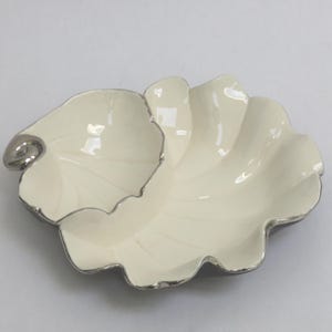 May include: Cream-colored, leaf-shaped decorative dish with a silver-colored trim. The dish has a glossy finish and a unique, organic design. The dish is ideal for serving small snacks or as a decorative accent piece.