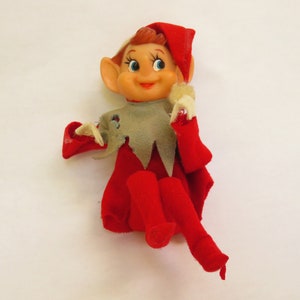 May include: A vintage elf doll with a red felt outfit, including a hat, cape, and tights. The doll has a plastic face with blue eyes, a smiling expression, and red hair. The elf's collar is gray and tattered.