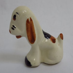May include: A ceramic dog figurine in a sitting position. The dog is primarily white with brown ears, black and brown spots, and black accents around the eyes and nose. The figurine has a glossy finish.
