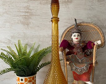 May include: Tall amber glass decanter with a textured, diamond-patterned body and a long, pointed stopper. A decorative plant in a retro floral pot and a seated puppet are also in the image. The decanter is a vintage home decor item.