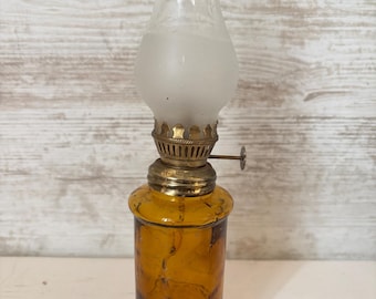 May include: A small amber glass oil lamp with a frosted glass chimney and a gold-colored metal top. The lamp has a decorative design on the glass.