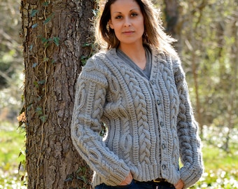 May include: A gray cable knit cardigan with a v-neck and button closure. The cardigan is worn by a person standing in a forest setting.