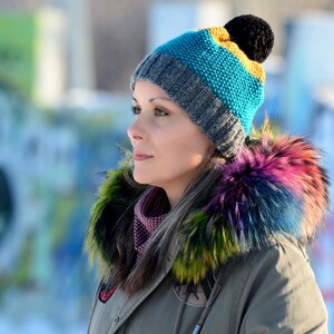 May include: A woman wearing a grey, blue and yellow knitted hat with a black pom-pom. The hat has a colourful fur trim.