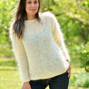 May include: A white fuzzy mohair sweater with a round neckline. The sweater is loose fitting and has long sleeves. The model is wearing the sweater with blue jeans.
